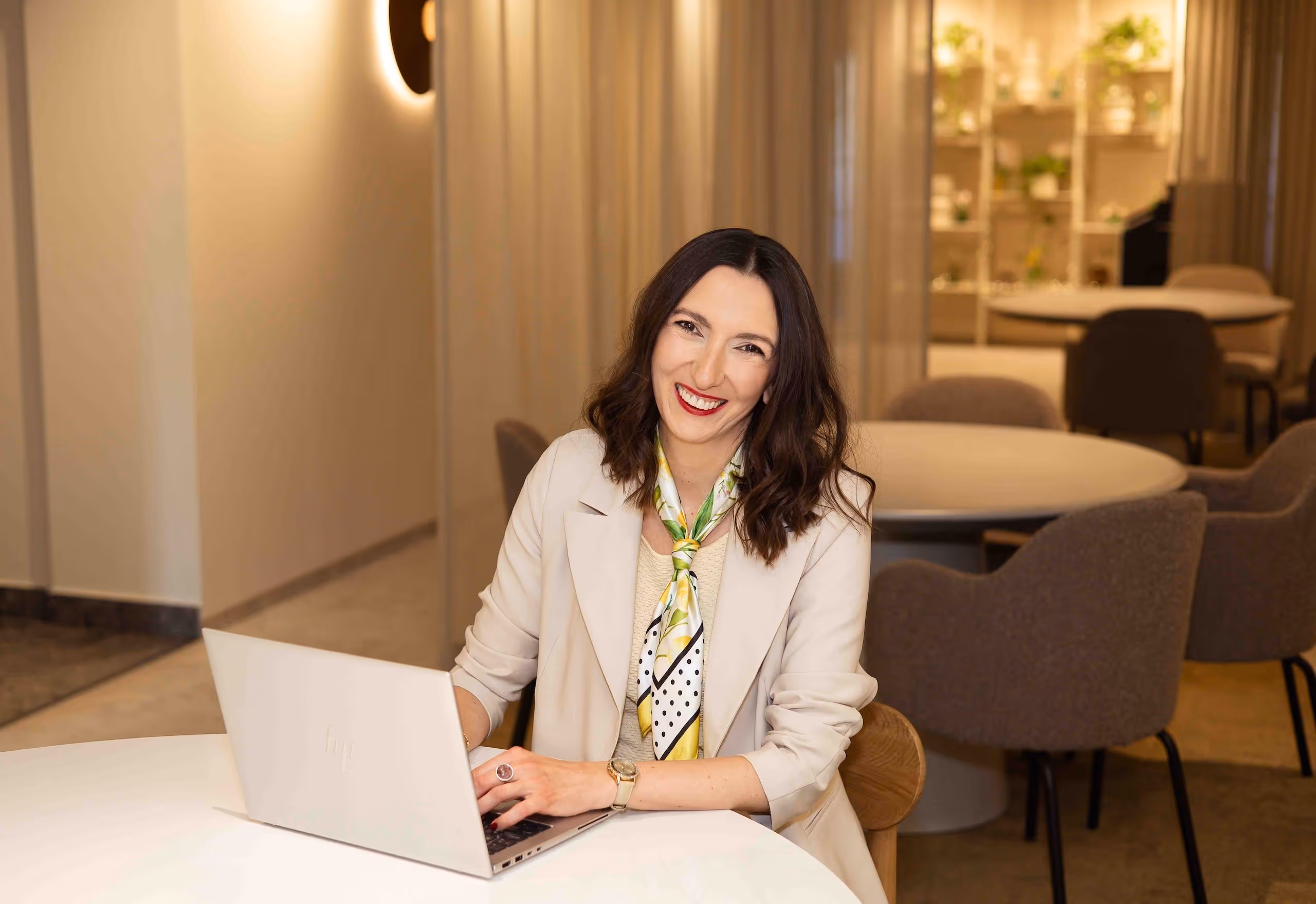 Alicja Dworowska, certyfikowana coach HeartMath®, uśmiecha się siedząc przy stole z laptopem, ubrana w jasną marynarkę i apaszkę.
