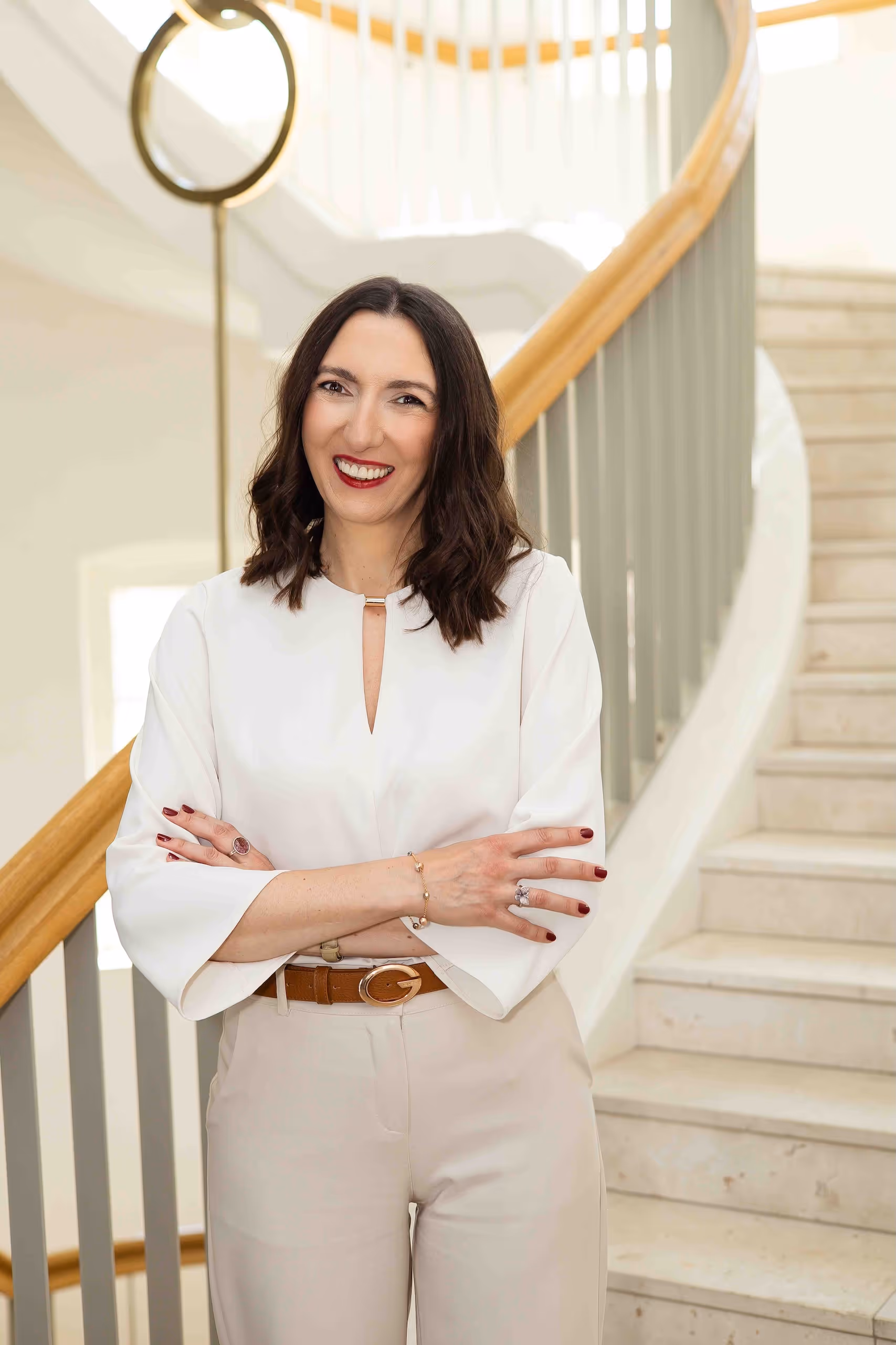Alicja Dworowska, certyfikowana coach HeartMath® i była CFO EMEA, stoi pewnie ze skrzyżowanymi rękami na tle eleganckiej marmurowej klatki schodowej.