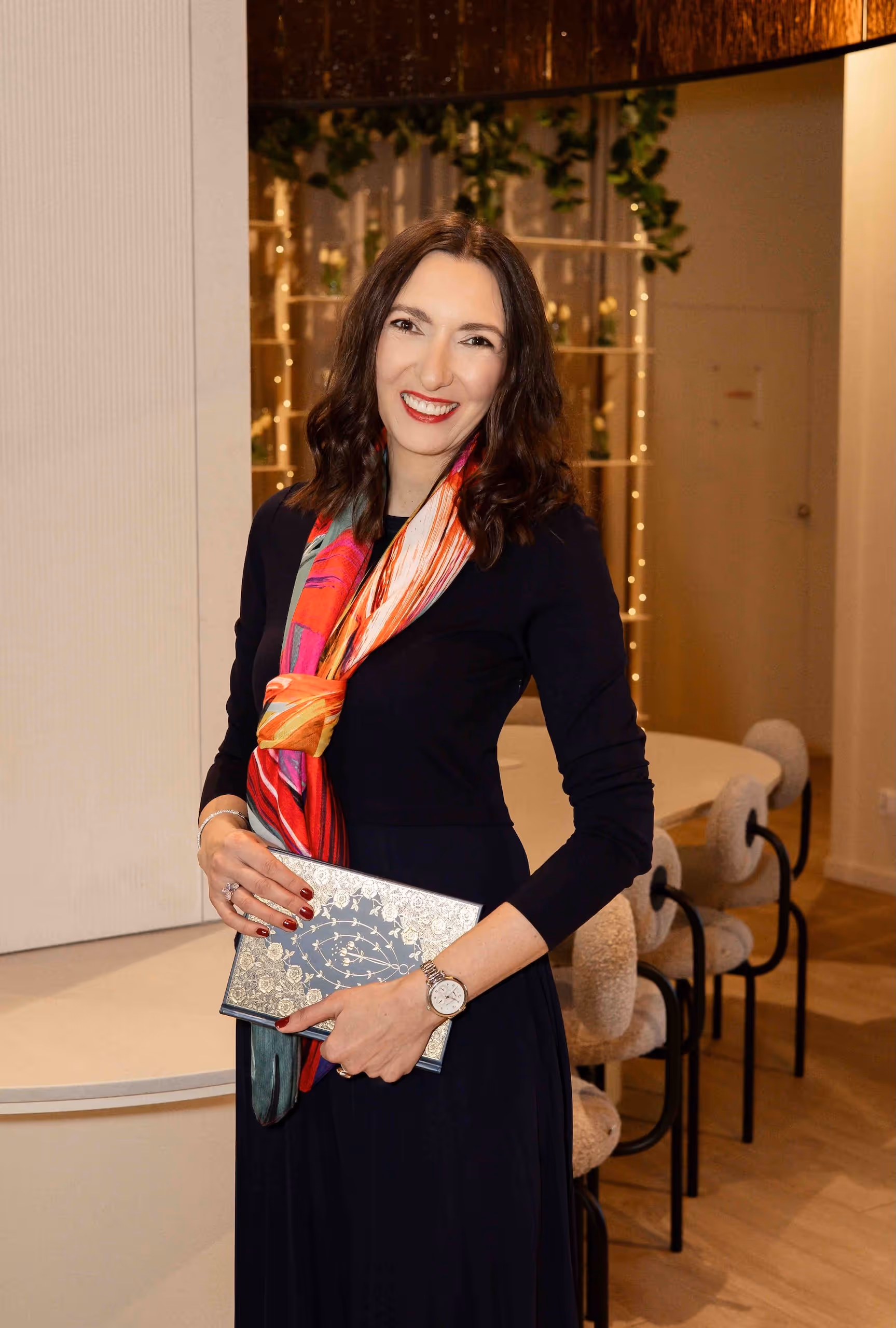 Uśmiechnięta Alicja Dworowska w czarnej sukni i kolorowym szalu trzyma ozdobną teczkę w eleganckim wnętrzu.